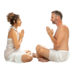 Ein Paar sitzt sich im Schneidersitz gegenüber, die Hände in Gebetshaltung, symbolisierend Achtsamkeit und Verbindung in der Tantra Massage und Meditation.